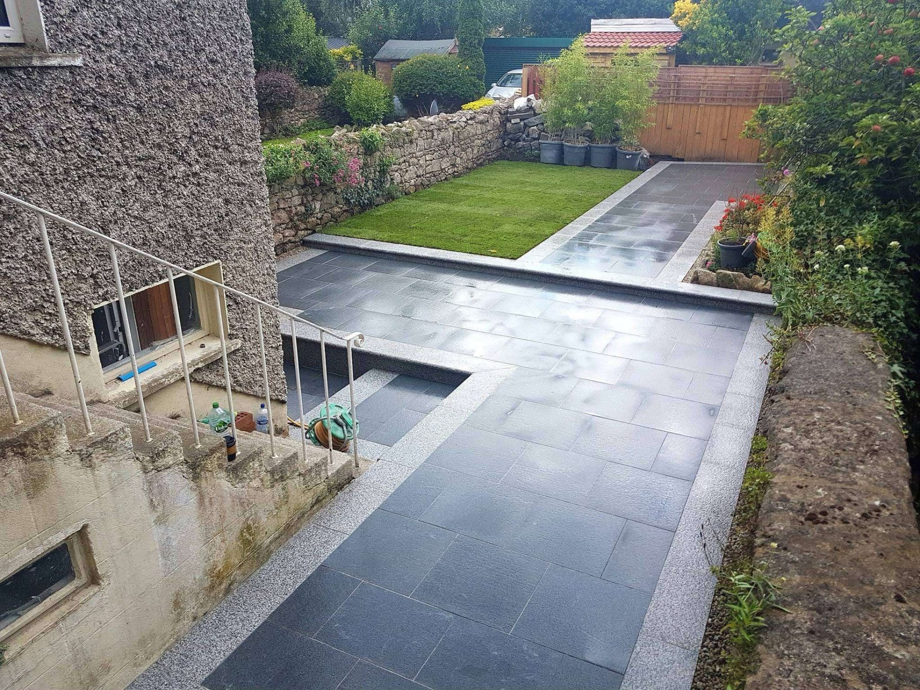 The same patio after landscaping — charcoal paving slabs, a new lawn and bamboo planters beside a stone wall