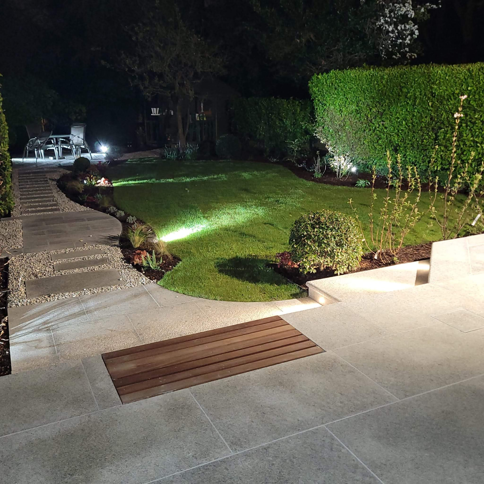 The same garden after landscaping — an evening view with lit lawns, granite steps, planted borders and pale stone paving